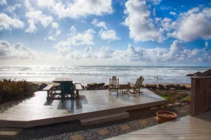 The image contains a beautiful outdoor uncovered wooden deck, facing the sea. On the Deck there is a table and some chairs to enjoy the scenery.